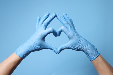 Doctor wearing medical gloves making heart gesture on light blue background, closeup