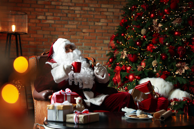 Santa Claus with hot drink and cookie near Christmas tree at home