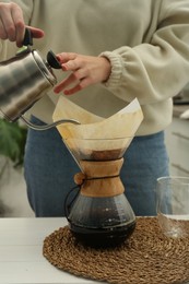 Photo of Woman pouring hot water into glass chemex coffeemaker with paper filter and coffee at table, closeup