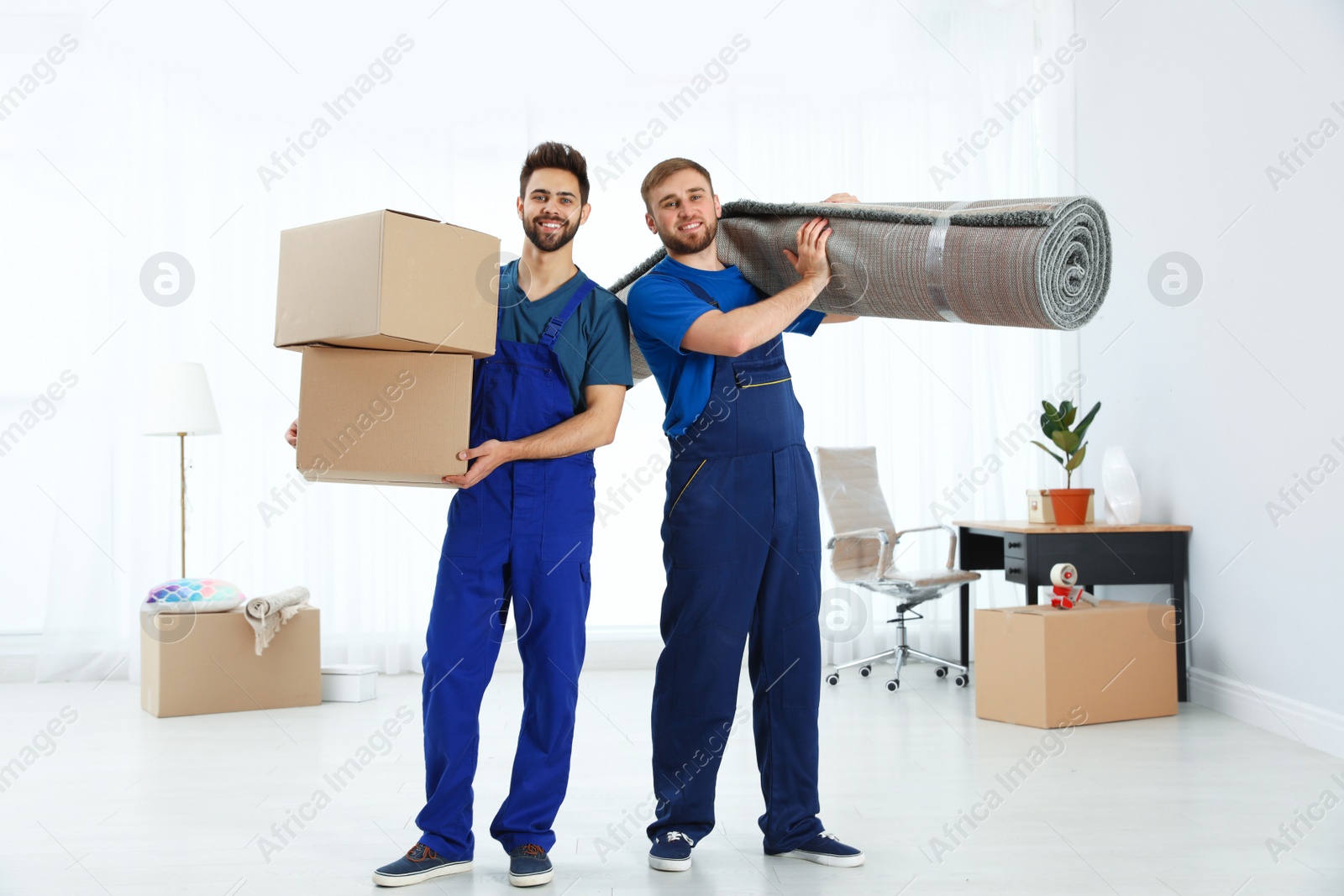 Photo of Young workers carrying rolled carpet and boxes in room. Moving service