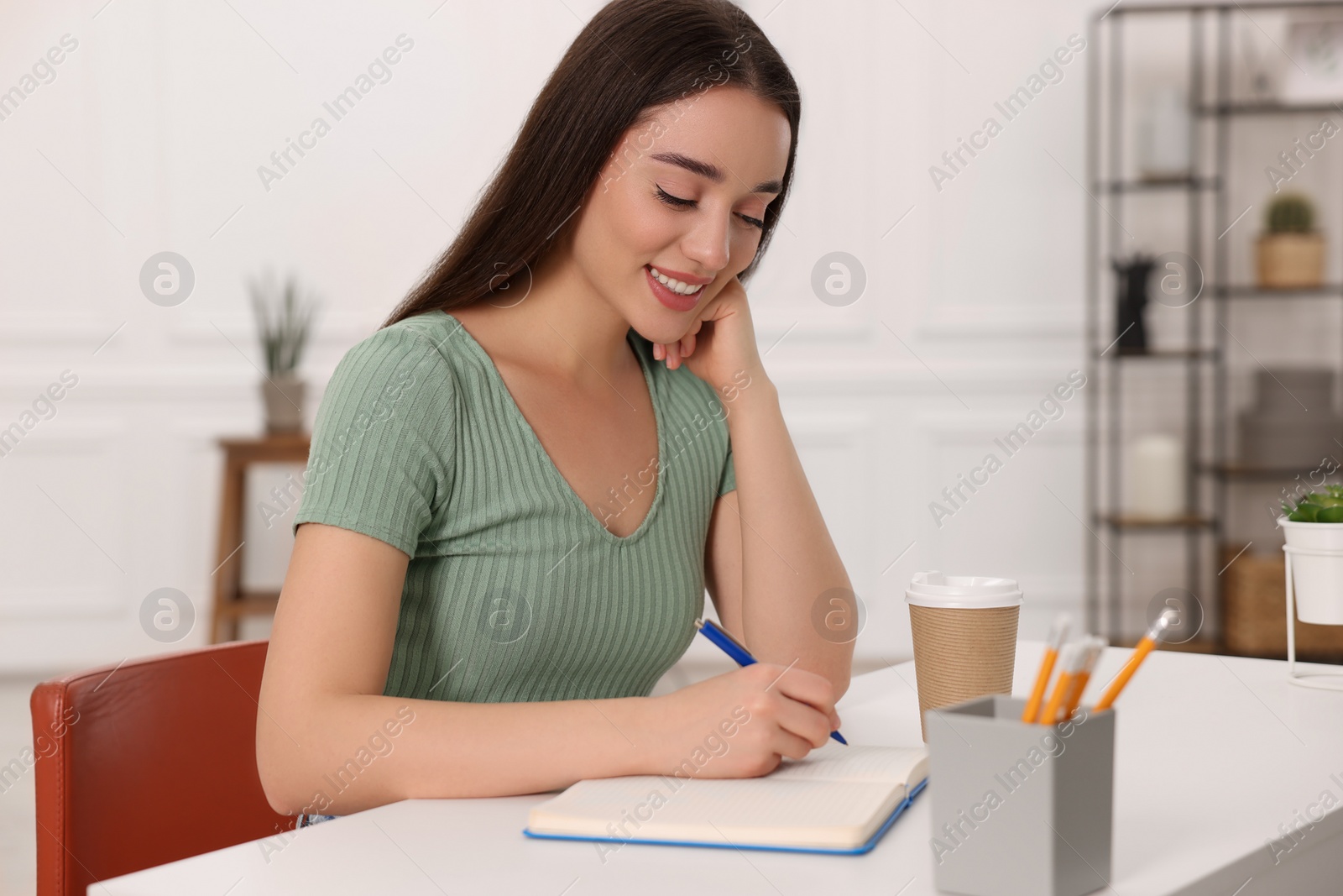 Photo of Young woman writing in notebook at white table indoors