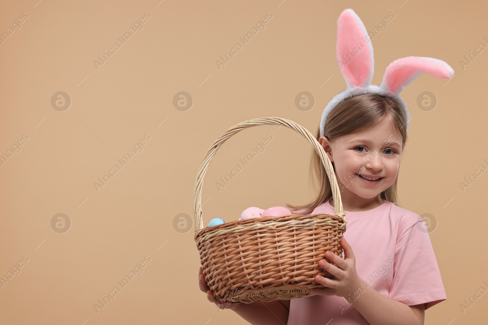 Photo of Easter celebration. Cute girl with bunny ears holding basket of painted eggs on beige background, space for text