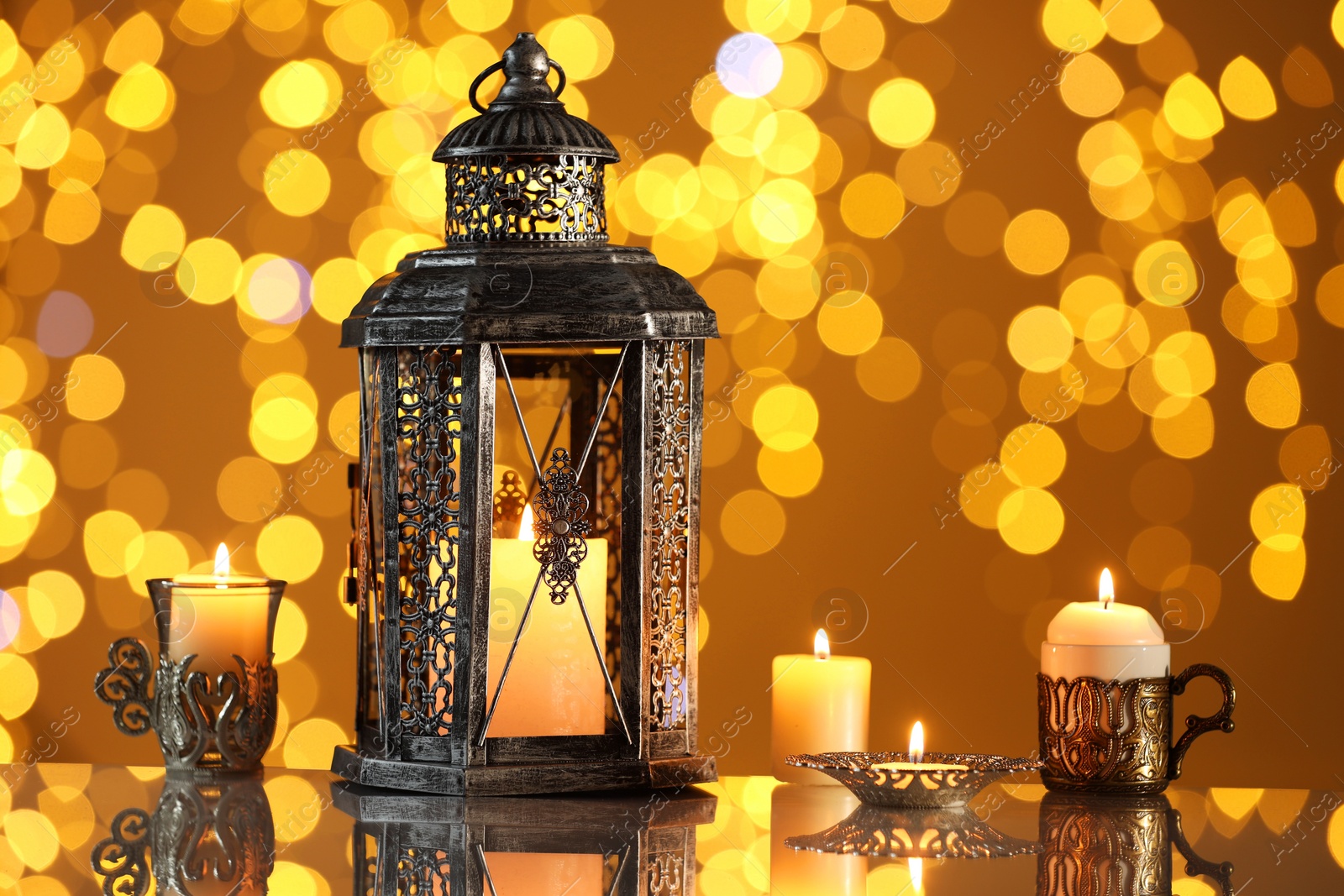 Photo of Arabic lantern and burning candles on mirror surface against blurred lights