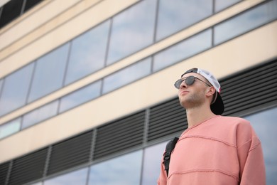 Photo of Handsome young man with stylish sunglasses and backpack near building outdoors, low angle view. Space for text