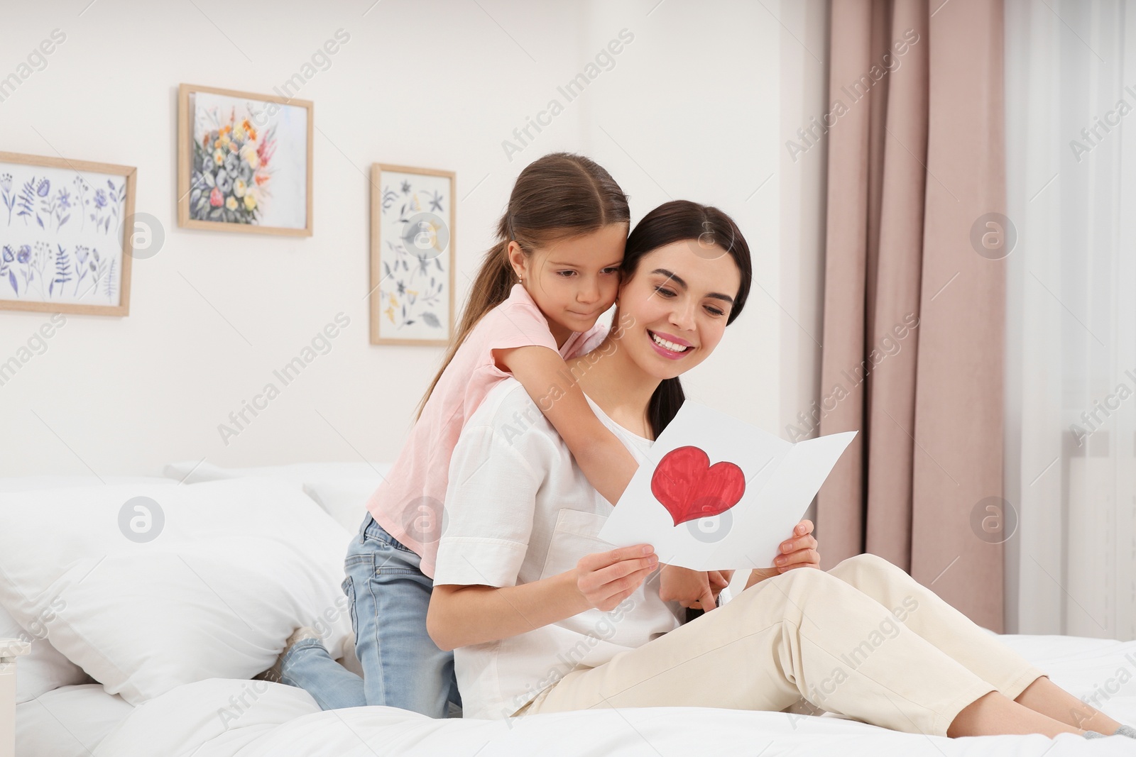 Photo of Happy woman with her daughter and handmade greeting card on bed at home. Mother's day celebration