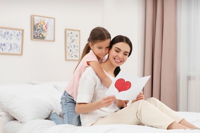 Photo of Happy woman with her daughter and handmade greeting card on bed at home. Mother's day celebration