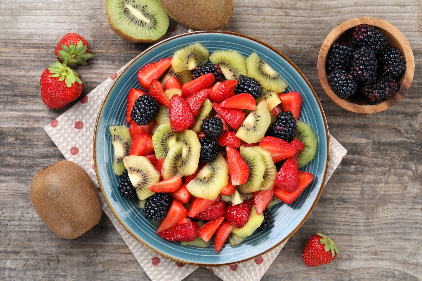 Photo of Plate of delicious fresh fruit salad and ingredients on wooden table, flat lay
