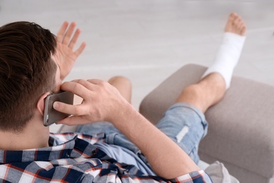 Photo of Young man with broken leg in cast talking on mobile phone at home