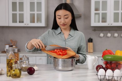 Photo of Cooking process. Beautiful woman adding cut bell pepper into pot in kitchen