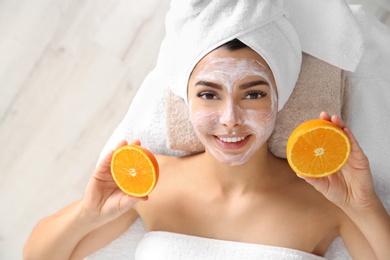 Pretty woman with rejuvenating facial mask holding sliced orange in spa salon, above view