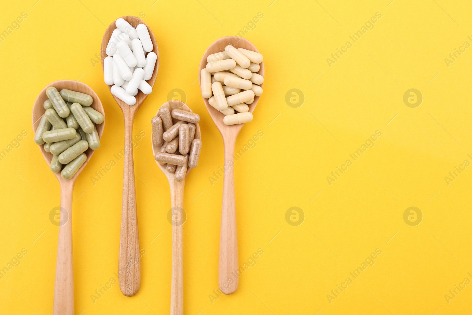 Photo of Different vitamin capsules in wooden spoons on yellow background, flat lay. Space for text