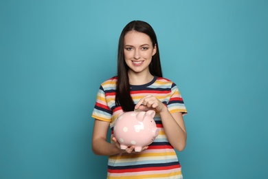 Young woman putting coin into piggy bank on color background