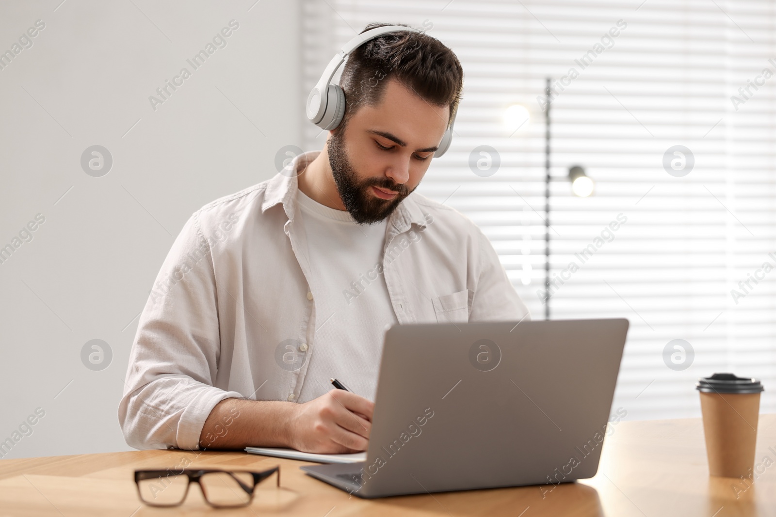 Photo of Young man in headphones writing down notes during webinar at table in room