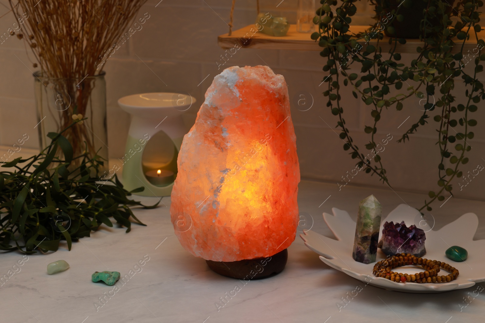 Photo of Himalayan salt lamp, crystals and oil diffuser on white table near brick wall