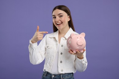 Excited woman pointing at piggy bank on purple background