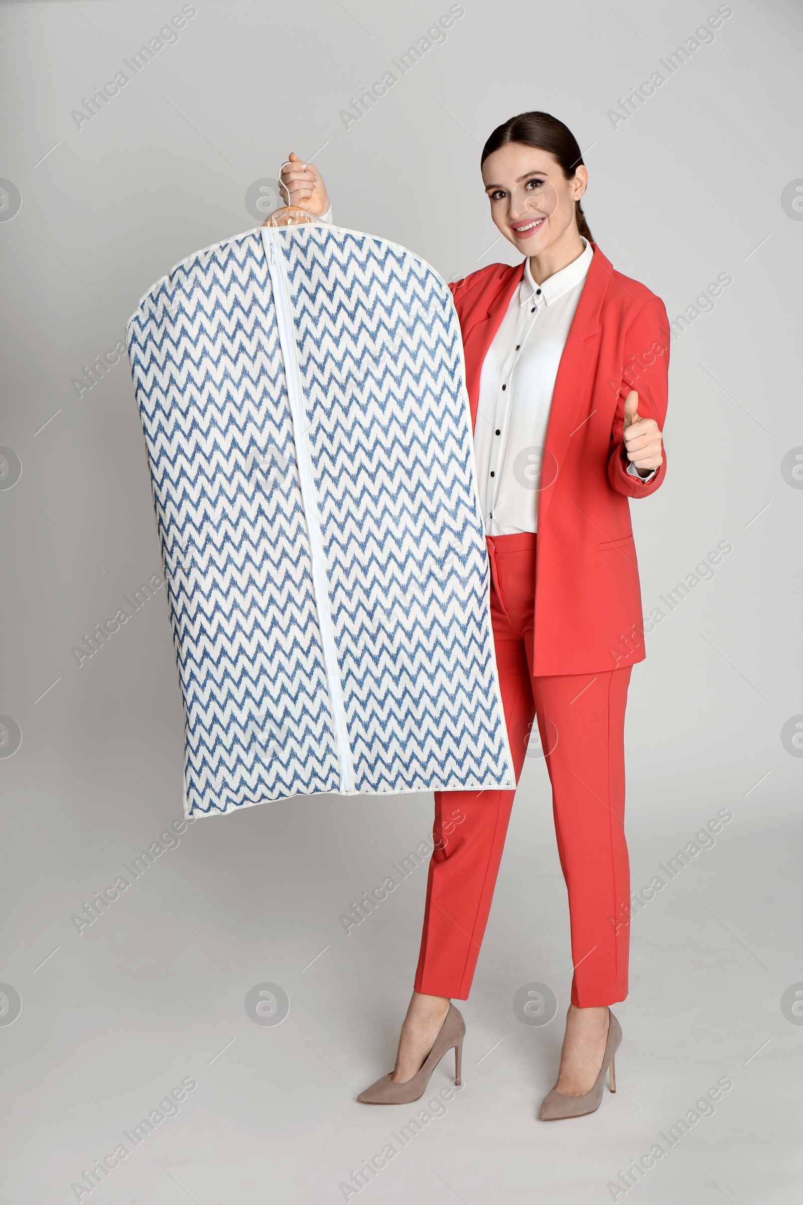 Photo of Young woman holding hanger with clothes in garment cover on light grey background. Dry-cleaning service