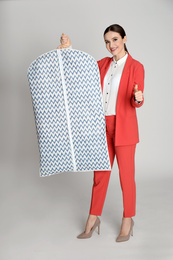 Photo of Young woman holding hanger with clothes in garment cover on light grey background. Dry-cleaning service