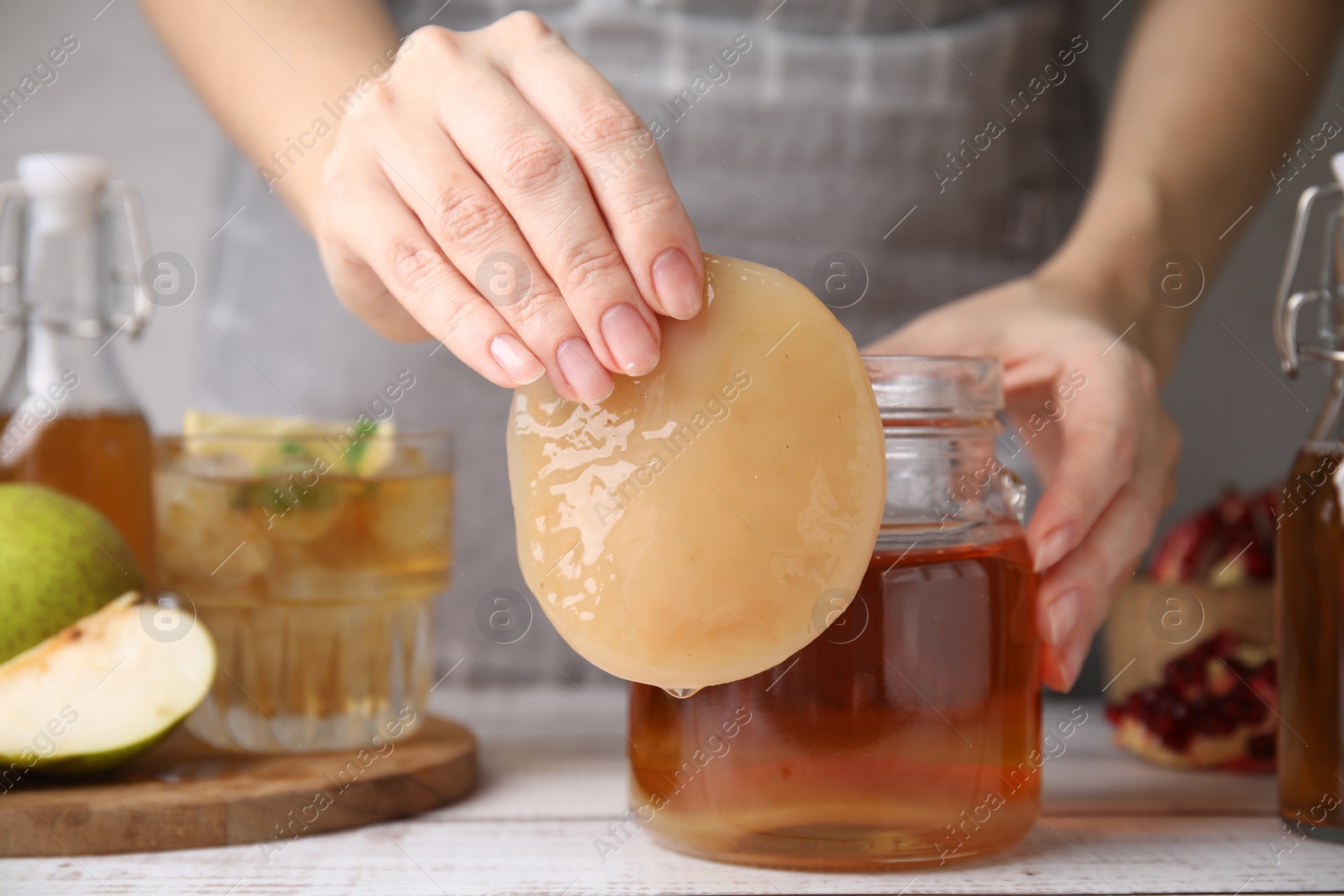 Photo of Woman putting Scoby fungus into jar with kombucha at white wooden table, closeup