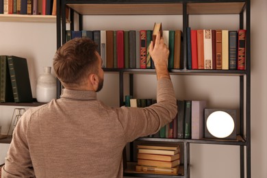 Photo of Young man choosing book on shelf in home library, back view