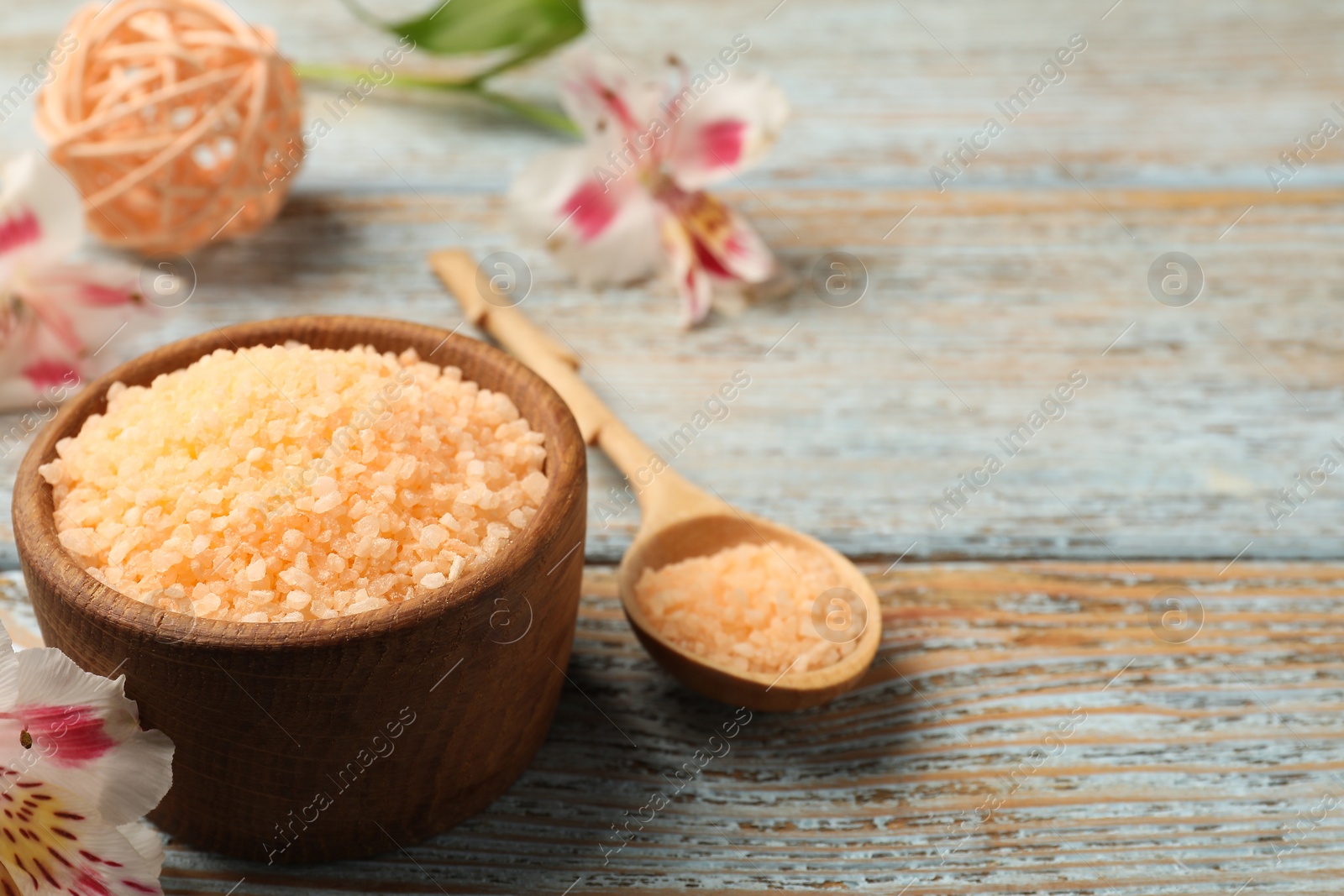 Photo of Orange sea salt in bowl, flowers and spoon on wooden table, closeup. Space for text