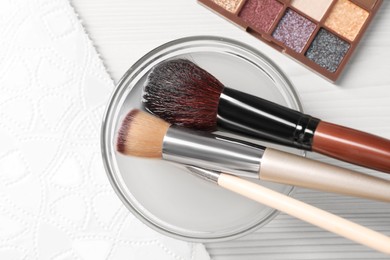 Photo of Cleaning makeup brushes in bowl with special liquid on white wooden table, flat lay