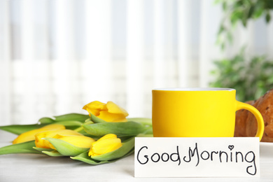 Croissant, coffee, beautiful flowers and card with words GOOD MORNING on white wooden table indoors