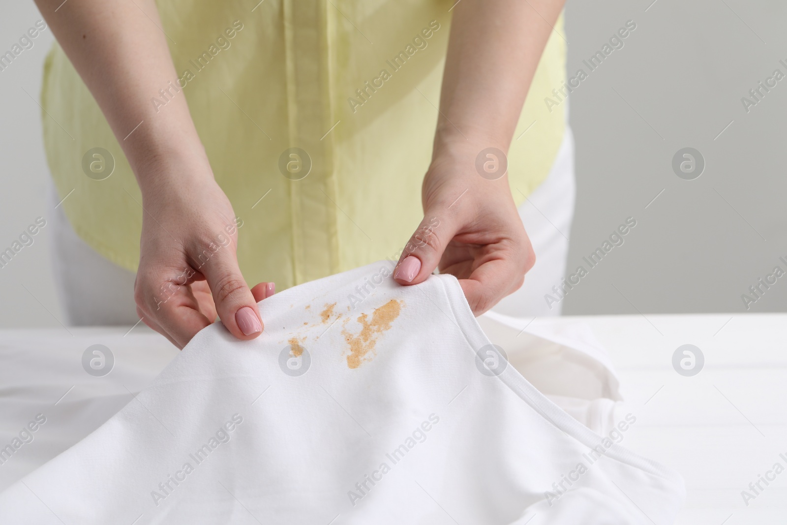 Photo of Woman holding shirt with stain at white table against light grey background, closeup