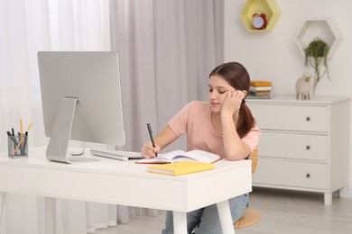 Photo of Tired girl writing in notepad while using computer at desk in room. Home workplace