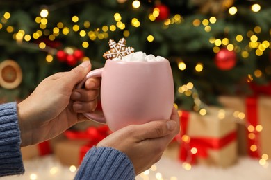 Photo of Woman holding cup of delicious cocoa with marshmallows and gingerbread cookie near Christmas tree at home, closeup