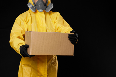 Photo of Man wearing chemical protective suit with cardboard box on black background, closeup. Prevention of virus spread