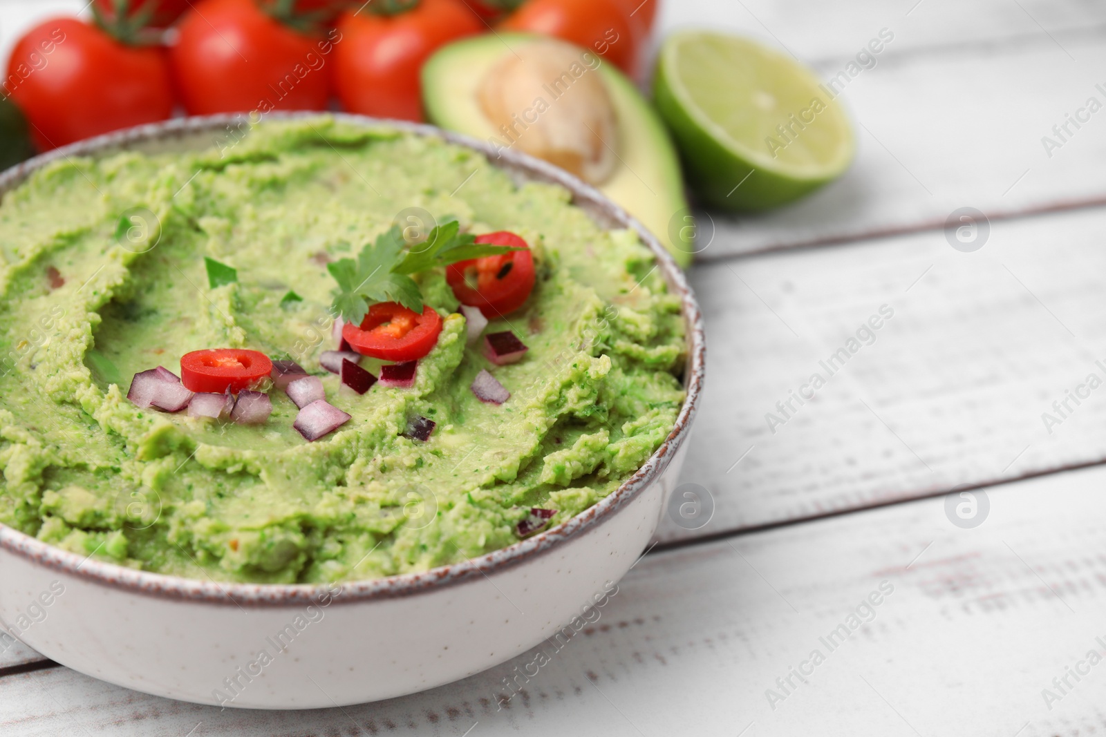 Photo of Bowl of delicious guacamole and ingredients on white wooden table, closeup. Space for text