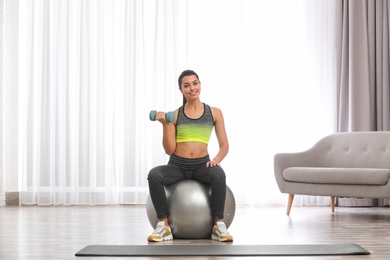 Young woman doing exercise with dumbbell on fitness ball at home
