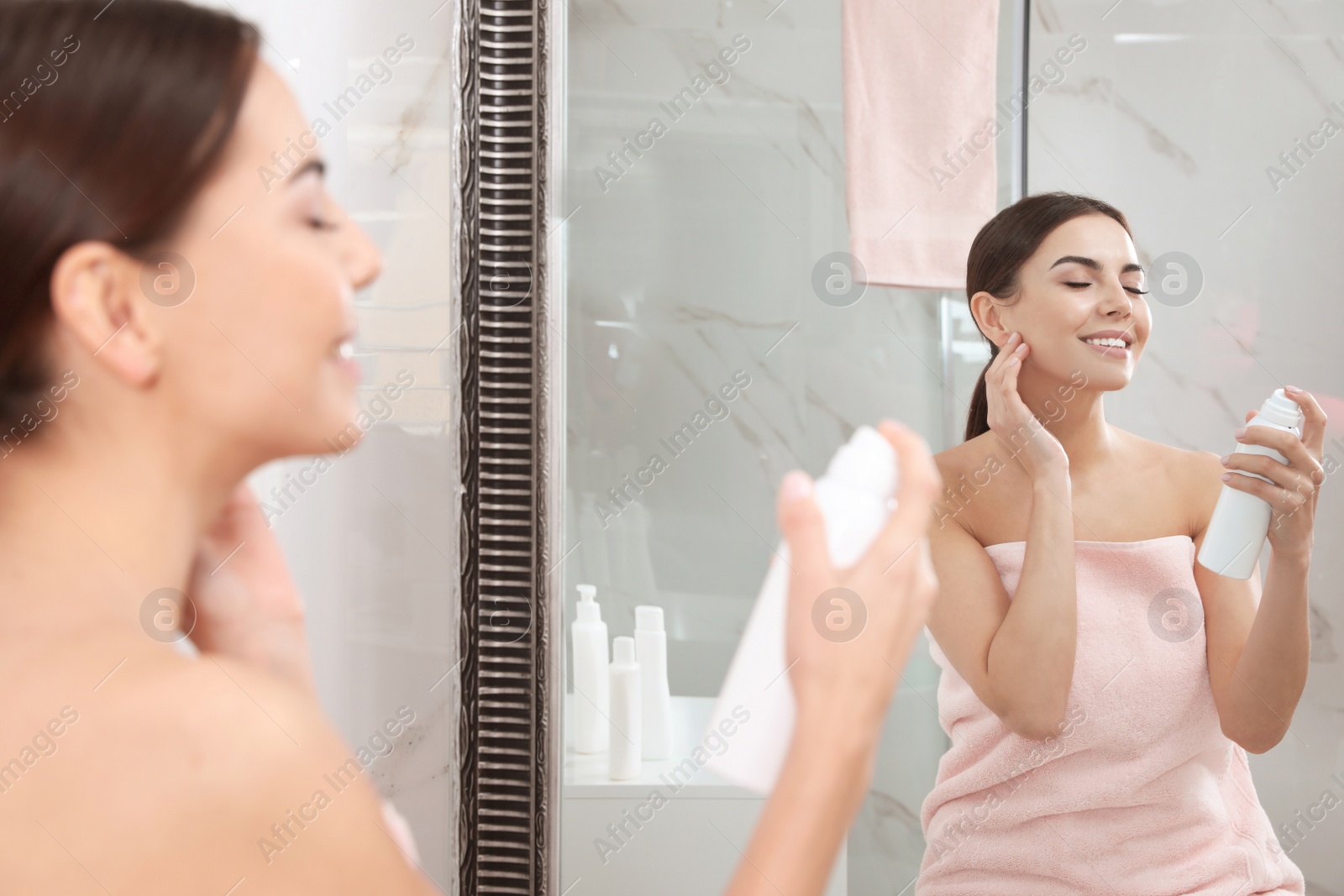 Photo of Young woman applying thermal water on face near mirror in bathroom. Cosmetic product