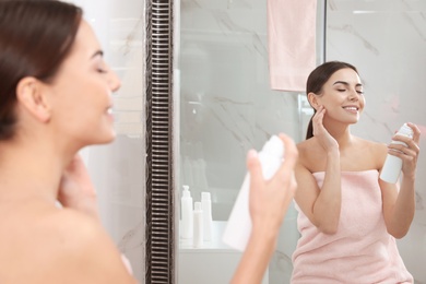 Young woman applying thermal water on face near mirror in bathroom. Cosmetic product