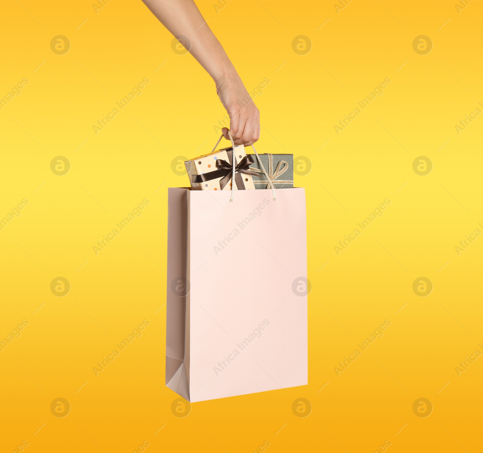 Photo of Woman holding paper shopping bag full of gift boxes on yellow background, closeup