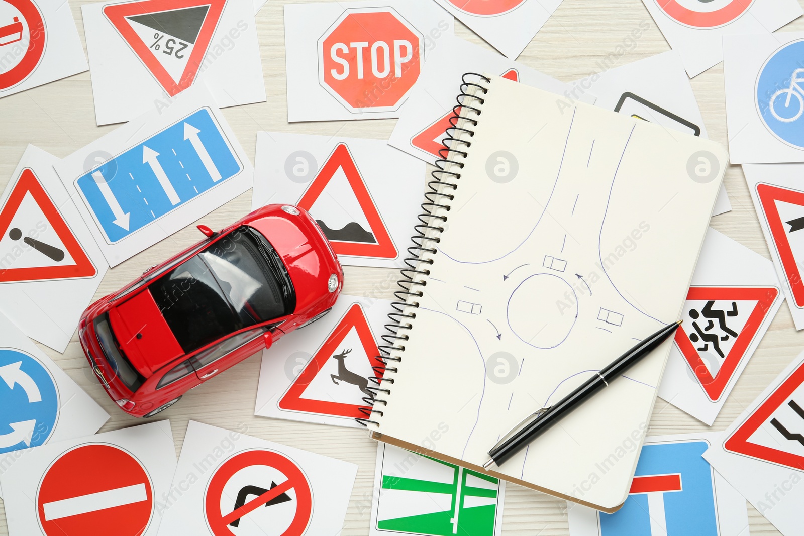 Photo of Many different road sign cards, notebook with sketch of roundabout and toy car on white wooden background, flat lay. Driving school