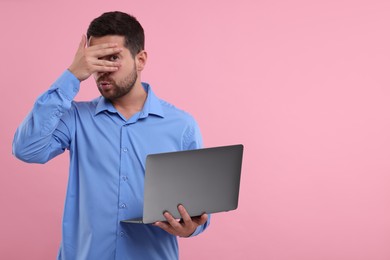 Photo of Embarrassed man covering face with hand and holding laptop on pink background. Space for text