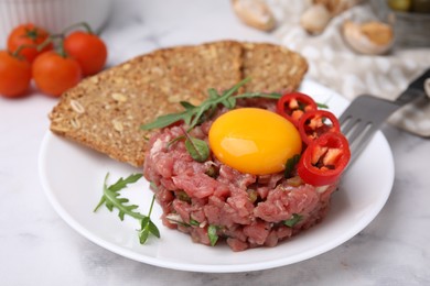 Photo of Tasty beef steak tartare served with yolk, pepper, bread and greens on white marble table