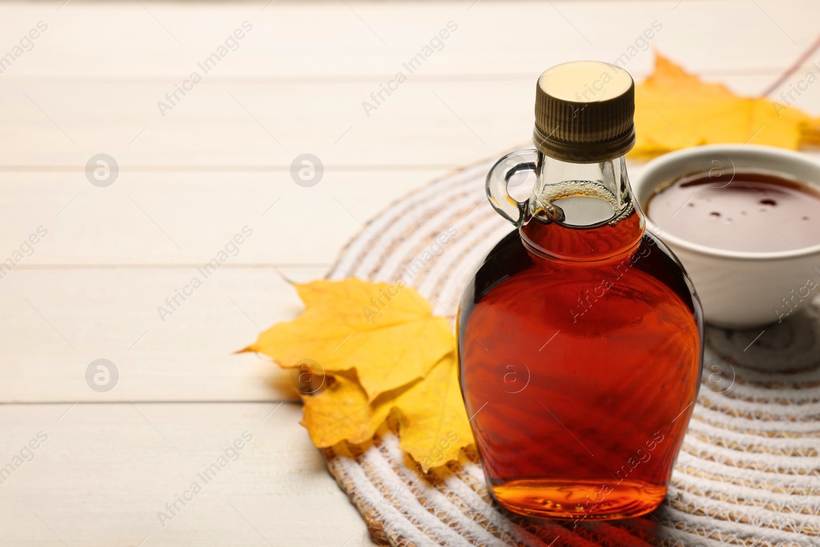 Photo of Bottle of tasty maple syrup, bowl and dry leaves on white wooden table, space for text