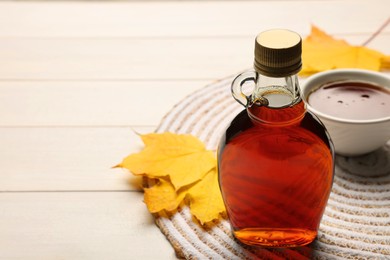 Photo of Bottle of tasty maple syrup, bowl and dry leaves on white wooden table, space for text