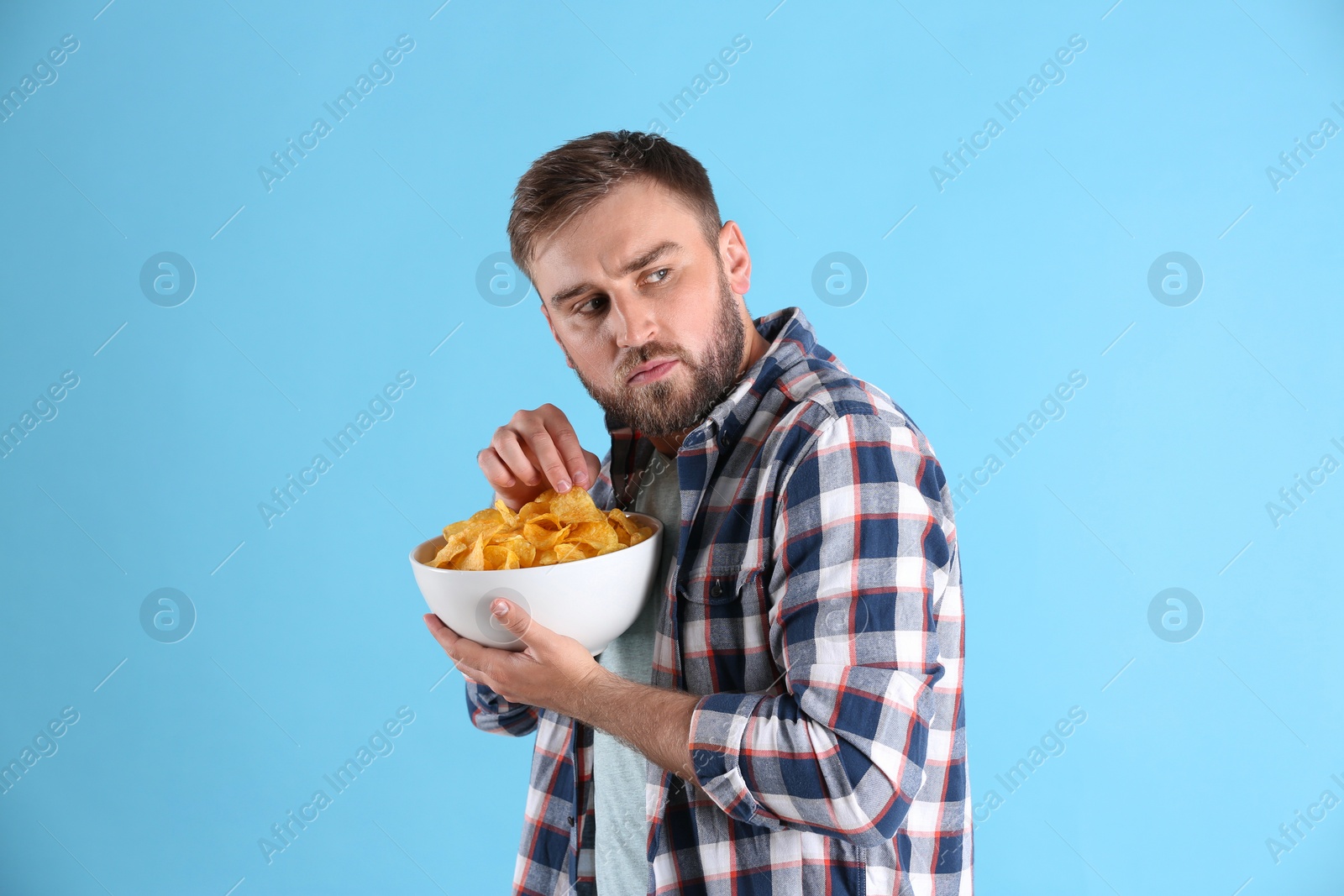 Photo of Greedy young man hiding bowl with chips on light blue background