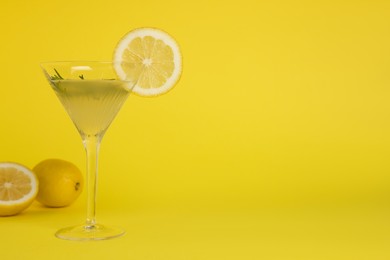Photo of Martini glass of refreshing cocktail with lemon slice, rosemary and fruits on yellow background, space for text