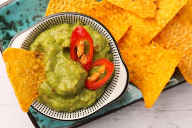 Bowl of delicious guacamole with chili pepper and nachos chips on white wooden table, top view