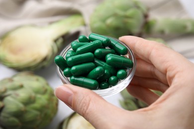 Photo of Woman holding dish of pills over table with fresh artichokes, closeup
