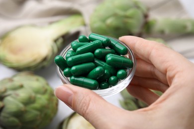 Woman holding dish of pills over table with fresh artichokes, closeup