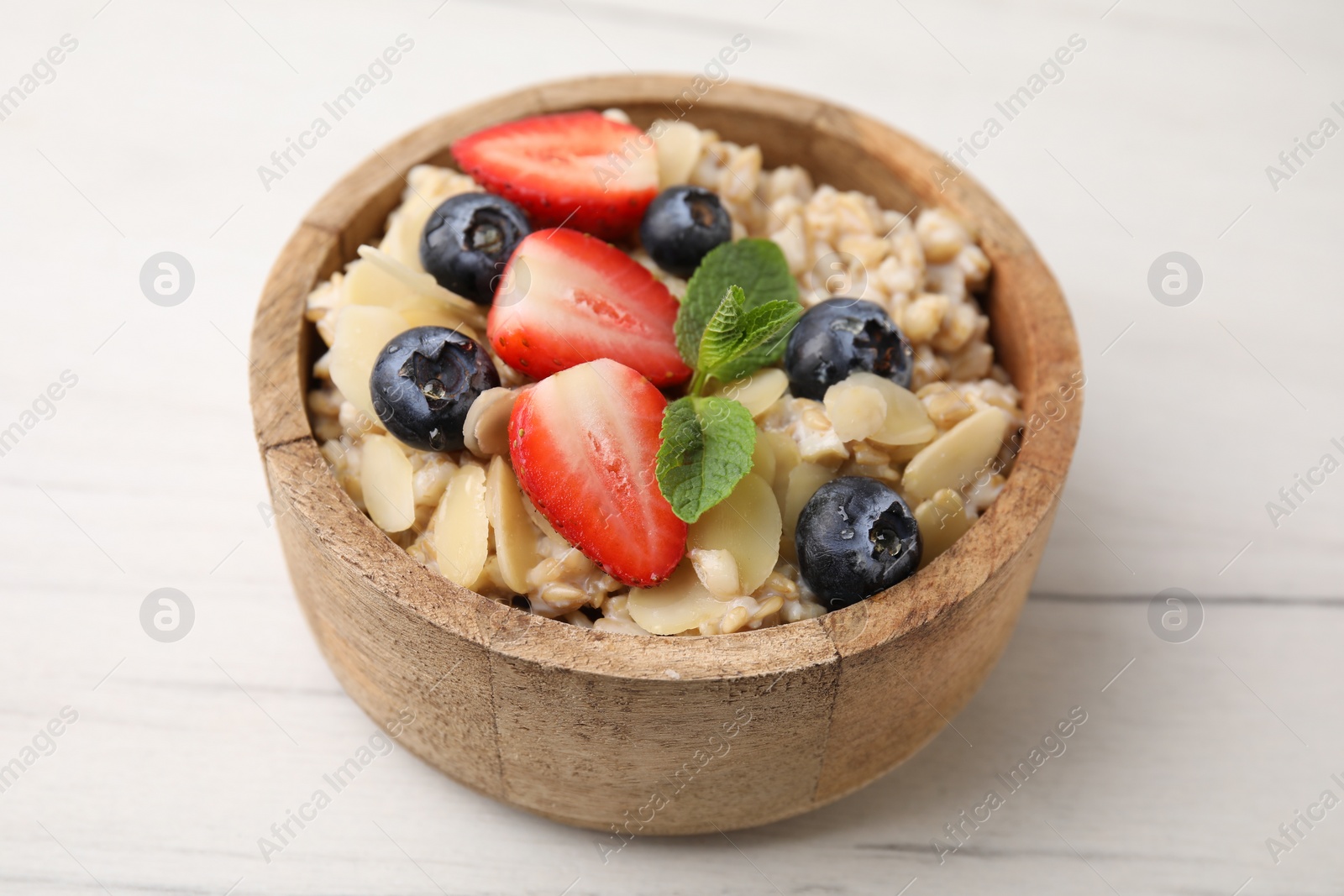 Photo of Tasty oatmeal with strawberries, blueberries and almond petals in bowl on white wooden table, closeup