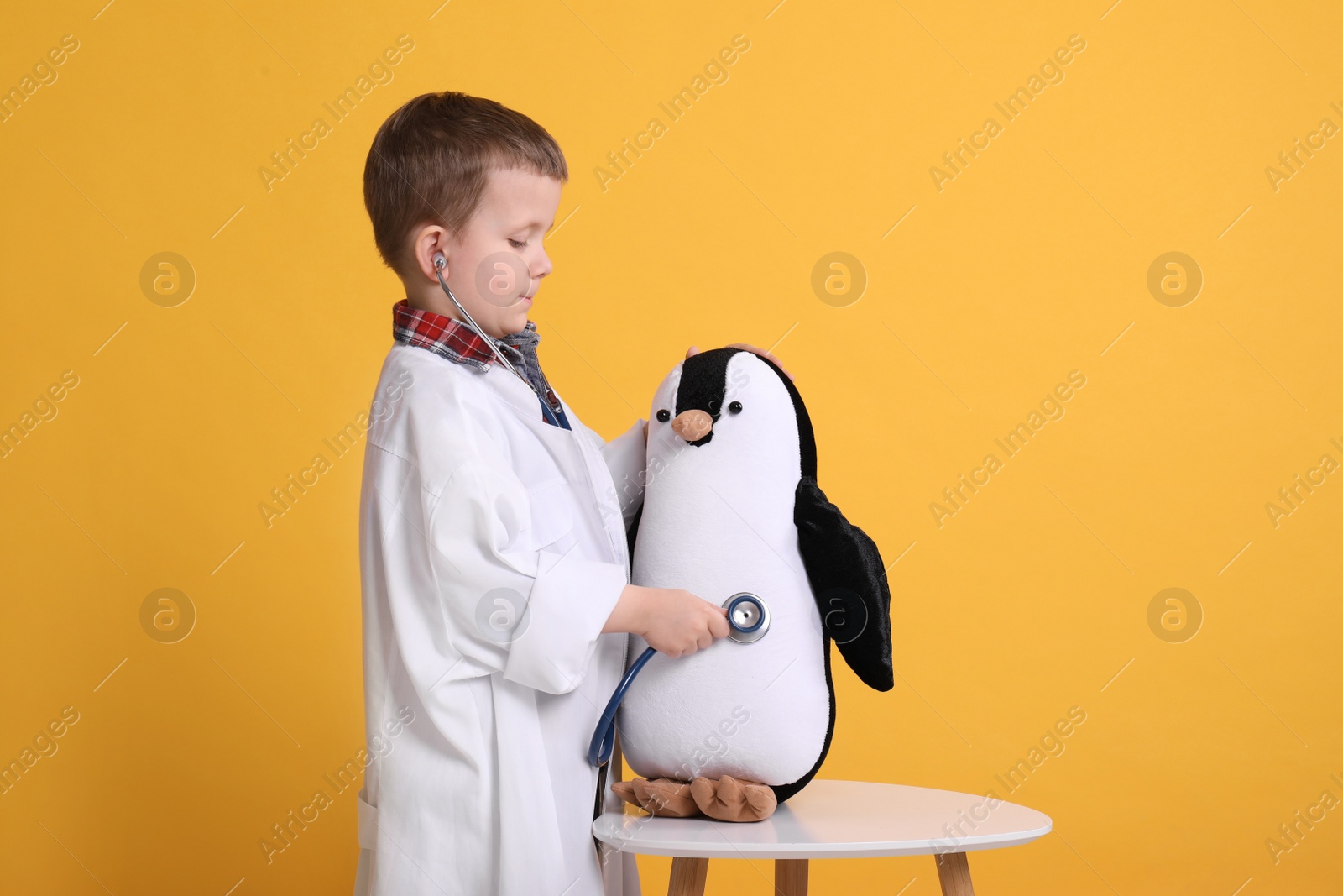 Photo of Cute little boy in pediatrician's uniform playing with stethoscope and toy penguin on yellow background