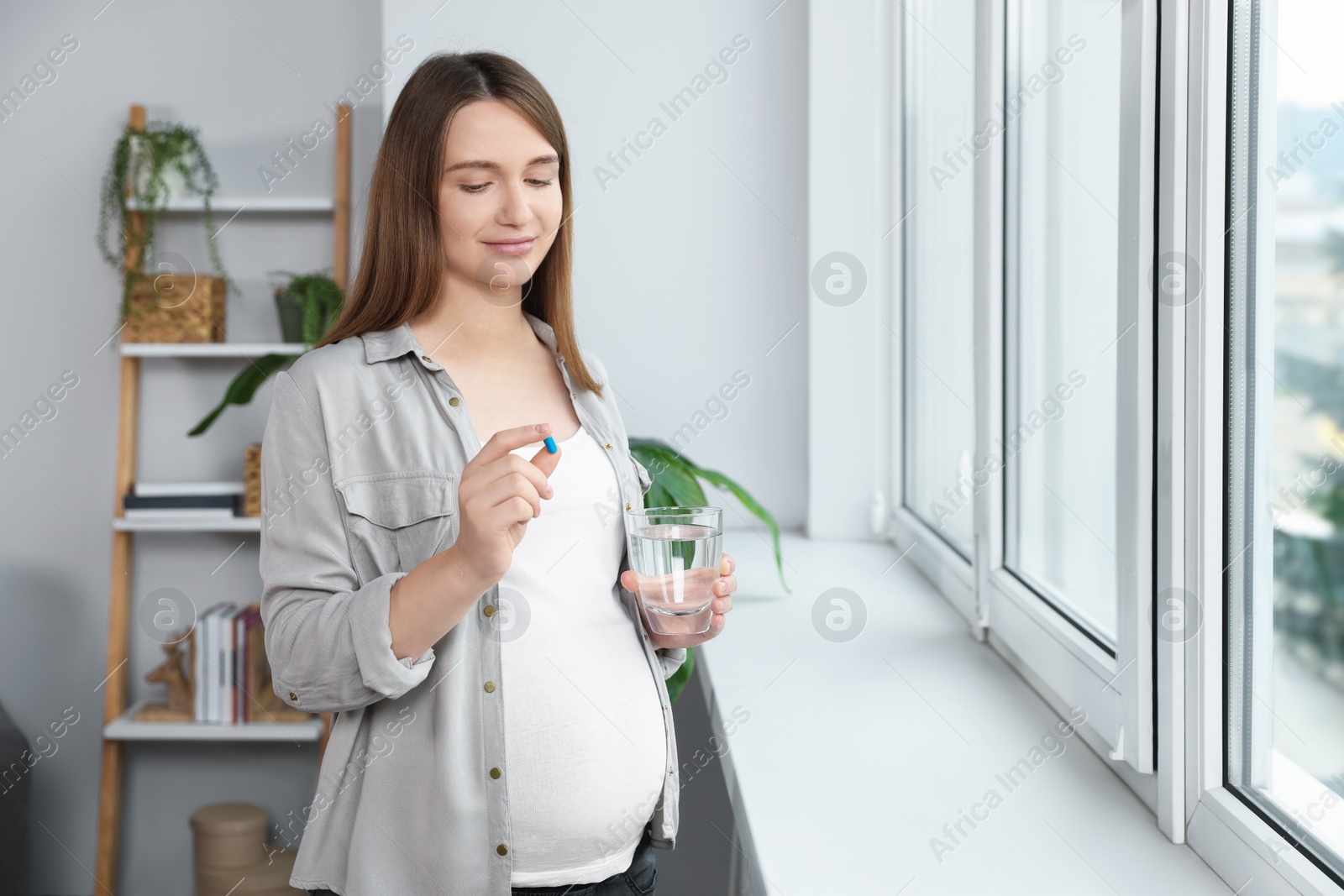 Photo of Beautiful pregnant woman holding pill and glass of water near window at home