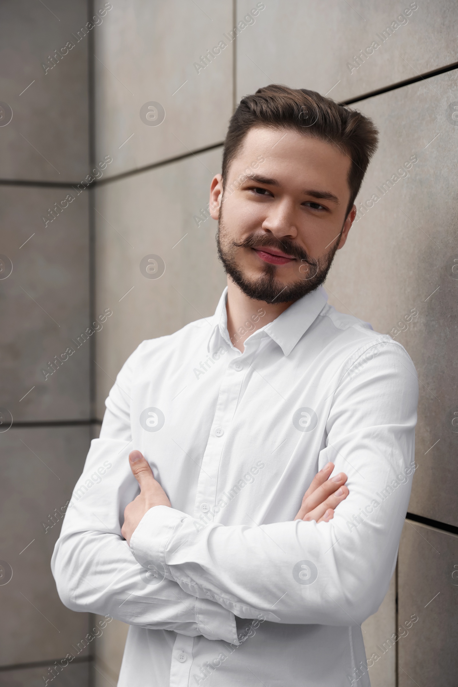 Photo of Smiling man in white shirt crossing his arms near light grey brick wall