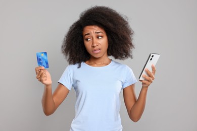 Confused woman with credit card and smartphone on grey background. Debt problem